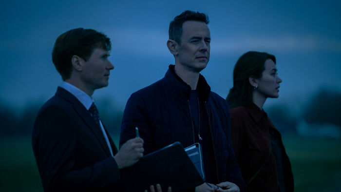 Three Silo Season 3 cast members, two men and a woman, stand outdoors in a dimly lit, somber setting. Three Silo Season 3 cast members, two men and a woman, stand outdoors in a dimly lit, somber setting.