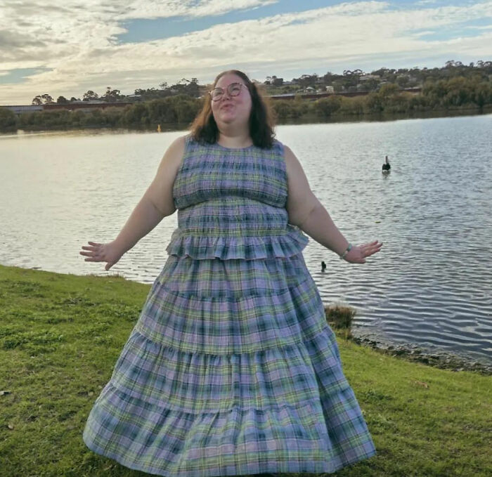 A plus-size influencer in a plaid tiered dress standing by a lake, expressing joy, amidst a reality check. A plus-size influencer in a plaid tiered dress standing by a lake, expressing joy, amidst a reality check.