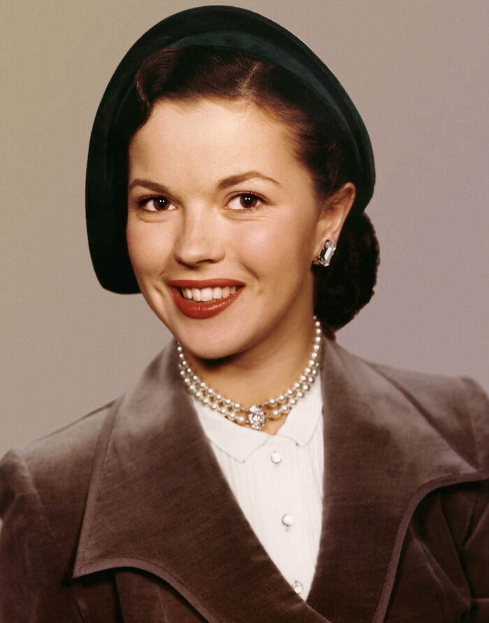 A portrait of a smiling woman wearing a dark hat, pearl necklace, and grey jacket. Fascinating things about the world.