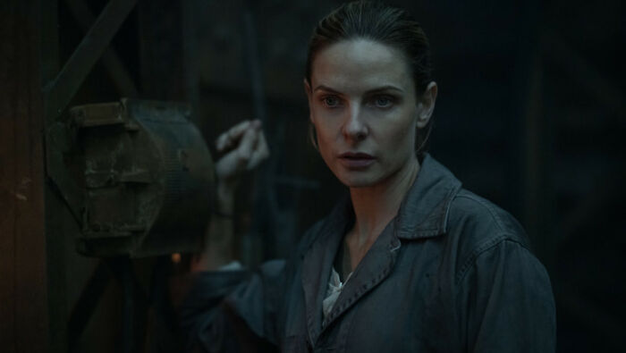 A woman with intense gaze, part of the Silo Season 3 cast, standing in a dimly lit, industrial setting. A woman with intense gaze, part of the Silo Season 3 cast, standing in a dimly lit, industrial setting.