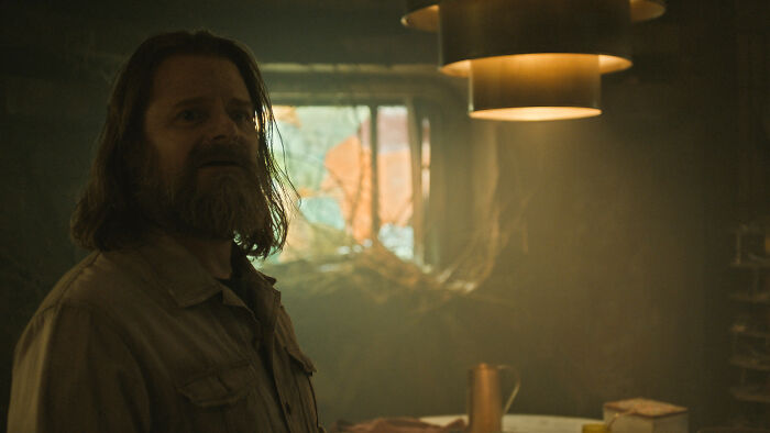 A man with long hair and a beard, a new character for Silo Season 3, looking worried in a cluttered room. A man with long hair and a beard, a new character for Silo Season 3, looking worried in a cluttered room.