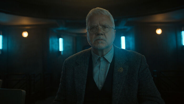 A thoughtful male Silo Season 3 cast character wearing glasses, a grey suit, and a dark vest, looking directly at the camera. A thoughtful male Silo Season 3 cast character wearing glasses, a grey suit, and a dark vest, looking directly at the camera.