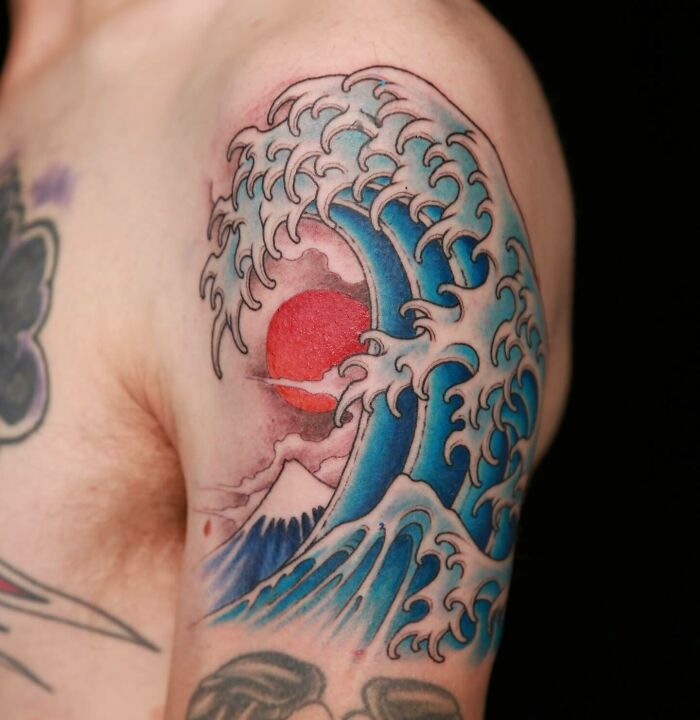 Shoulder tattoo art depicting a crashing wave with a red sun and mountain, a classic example of inspiring tattoo art.