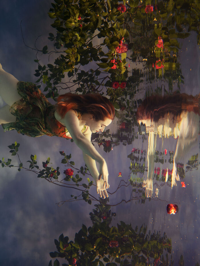 A woman with red hair creates surreal photos by submerging herself in water, surrounded by roses and their reflection.