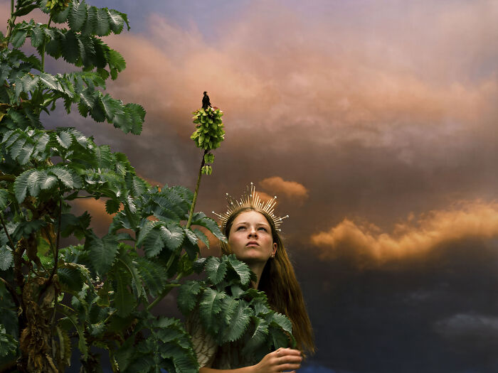 Surreal photo of a woman wearing a crown looking up at a dramatic sky, peeking from behind green leaves.