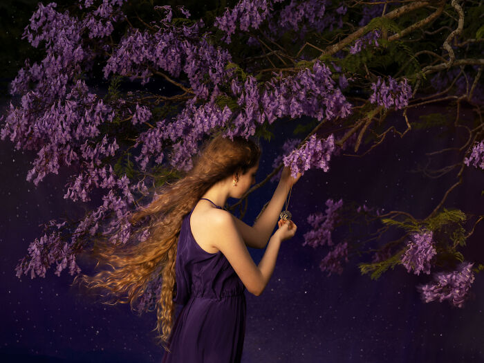 Surreal photo of a woman with long hair in a purple dress reaching for a branch with purple flowers against a starry background.