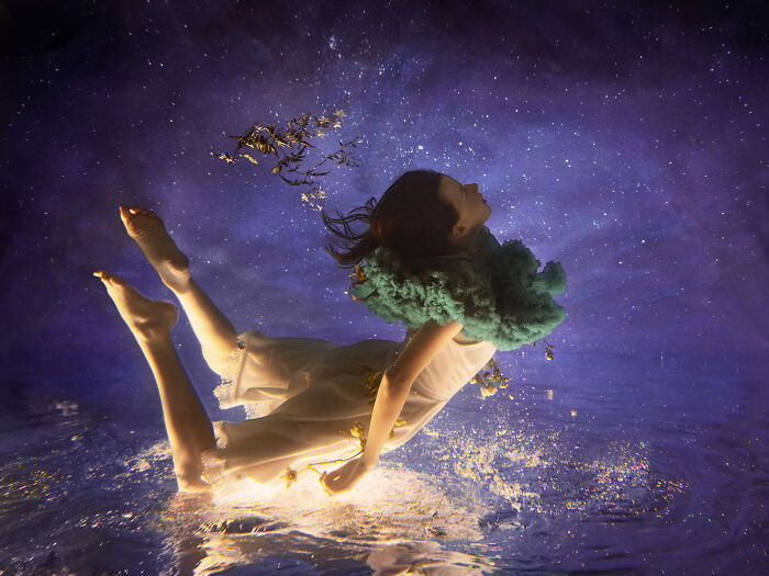 Surreal photo of a woman in a white dress with a green ruffled collar submerging in water under a starry sky.