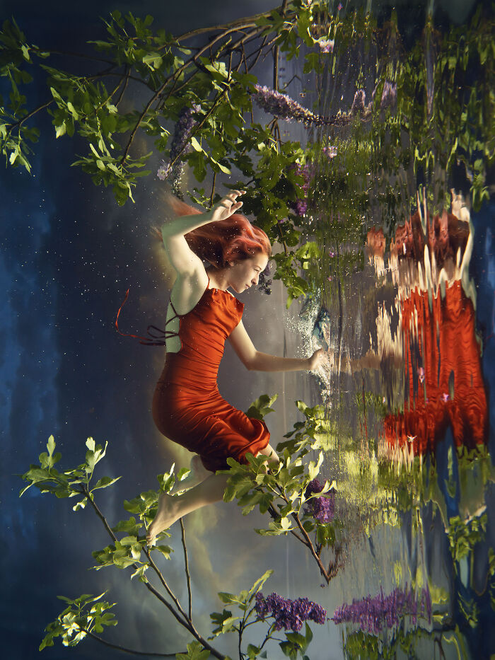 Surreal photo of a woman in a red dress submerging in water, with green leaves and purple flowers surrounding her.