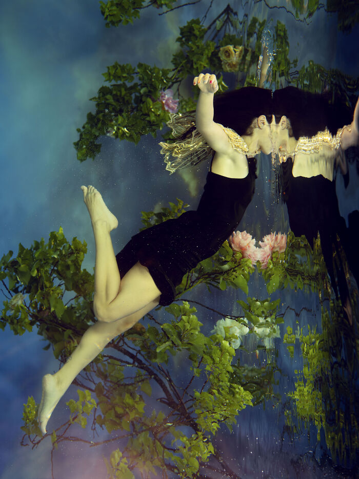 Surreal photo of a woman submerging in water, her reflection visible, surrounded by green leaves and pink flowers.