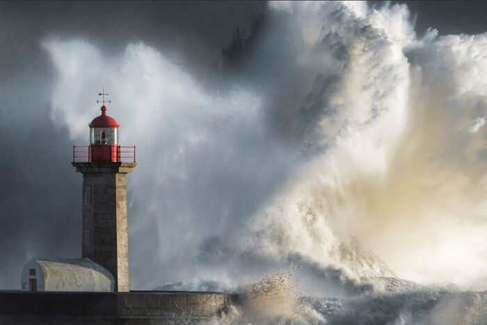 A dramatic color photo capturing the world: a lighthouse standing firm against a massive wave crashing during a storm.