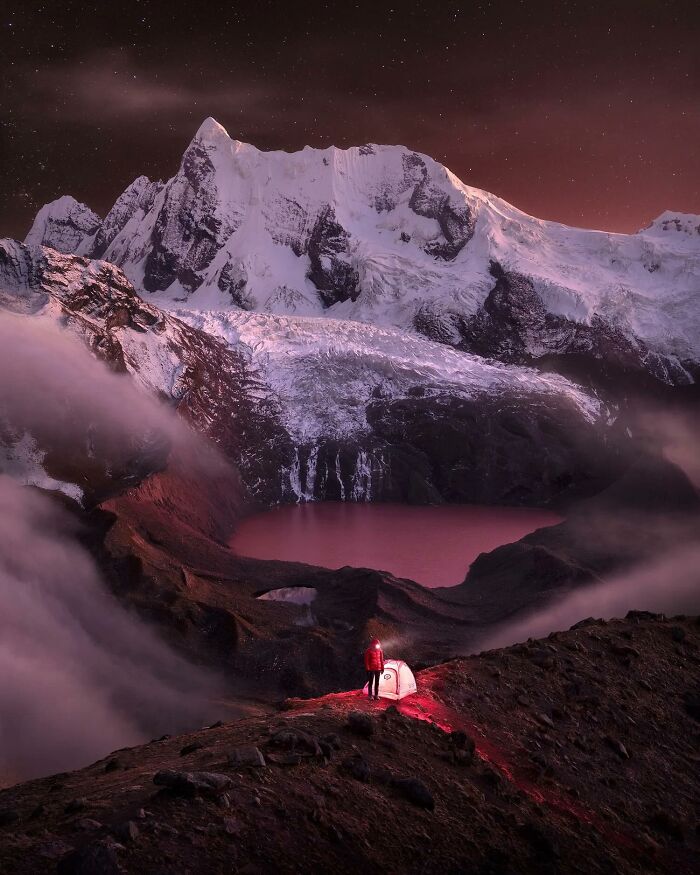 Remarkable color photo capturing the world: a lone camper by a tent, overlooking a pink lake and snowy mountains.