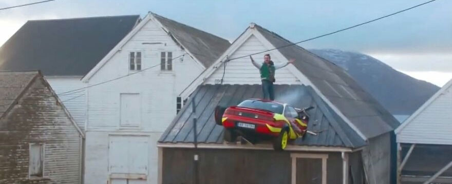 A red and yellow car perched precariously on a rooftop with a person standing nearby, a rough day behind the wheel.