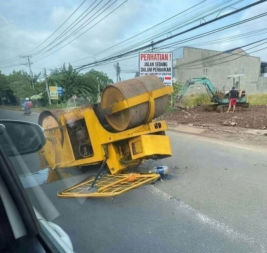 A yellow road roller overturned on its side in the middle of a road construction zone, a rough day behind the wheel.