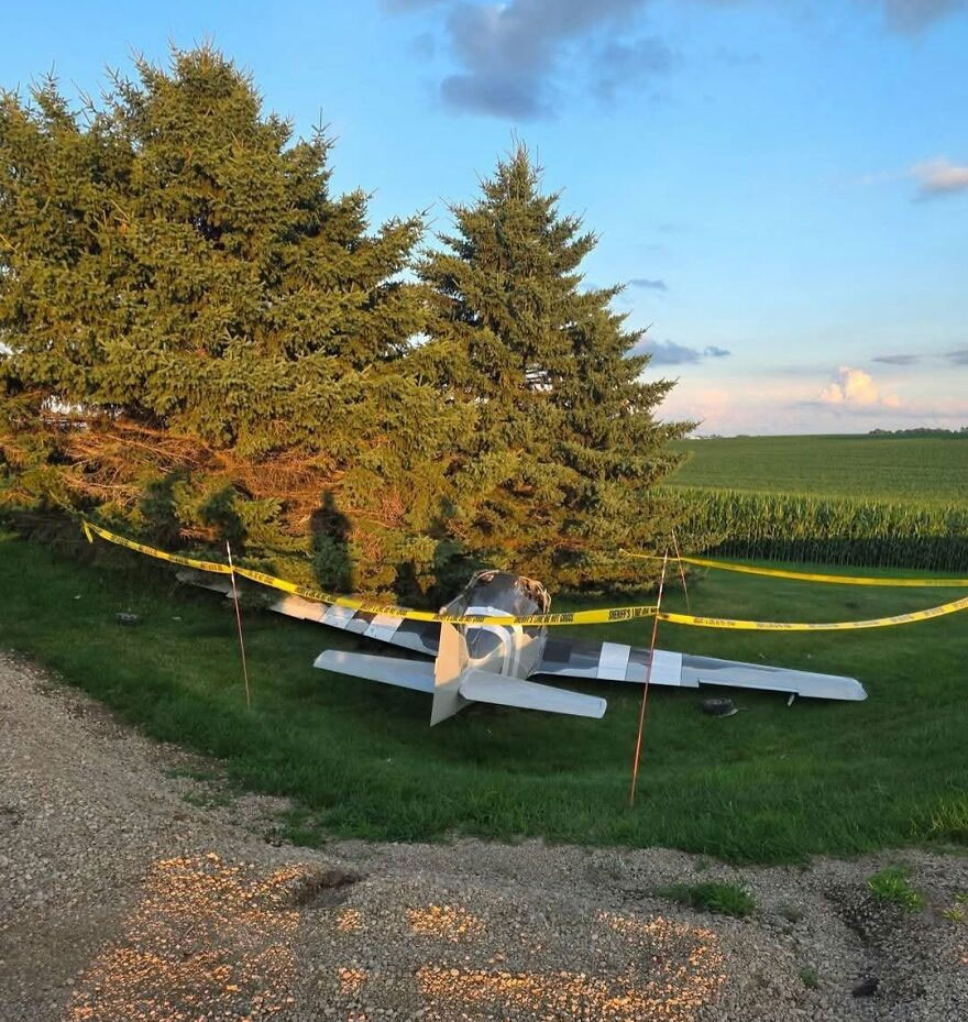 A small plane crash-landed next to trees in a field, marked with yellow caution tape, showing a rough day.