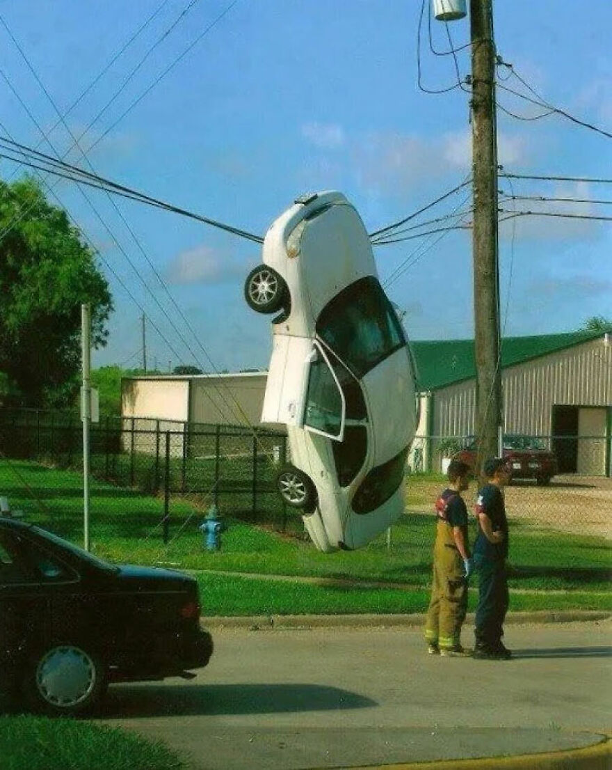 A white car suspended vertically in the air, entangled in power lines, as two men look on, showing a rough day behind the wheel.