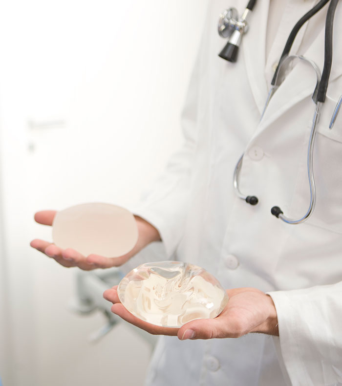 A doctor holding two breast implants, one opaque and one clear, demonstrating the difference in removed implants. A doctor holding two breast implants, one opaque and one clear, demonstrating the difference in removed implants.