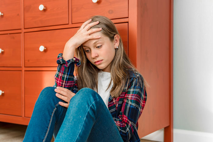 A teenage daughter looks upset, sitting by a dresser, after losing her college fund due to a family conflict. A teenage daughter looks upset, sitting by a dresser, after losing her college fund due to a family conflict.