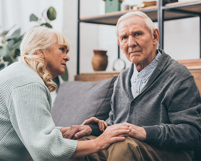 An elderly couple sitting on a couch, holding hands, with a doctor sharing warning symptoms. An elderly couple sitting on a couch, holding hands, with a doctor sharing warning symptoms.