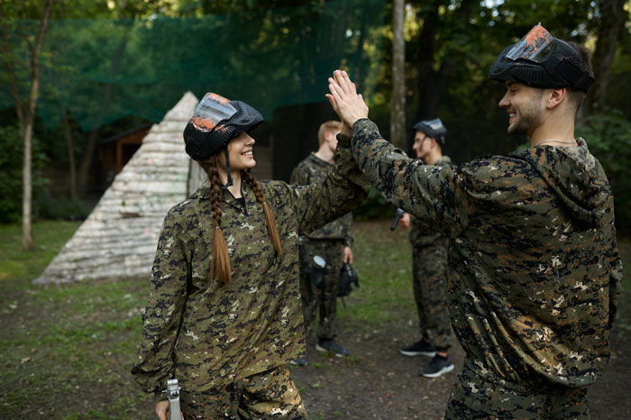 Two happy airsoft players in camo, a man and a woman, high-five each other while ignoring dress code. Two happy airsoft players in camo, a man and a woman, high-five each other while ignoring dress code.