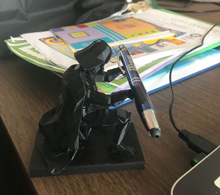 A black Darth Vader figurine, a 3D printing creation, holds a pen on a desk, an example of amazing 3D printing creations.