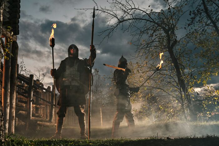Two armored figures holding torches and a spear in a misty, dark setting, evoking historical myths and ancient battles.