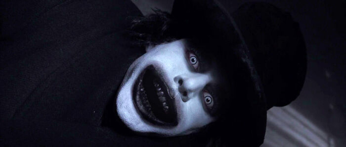 A person in a black hat with a white, grinning, terrifying face from a horror movie.