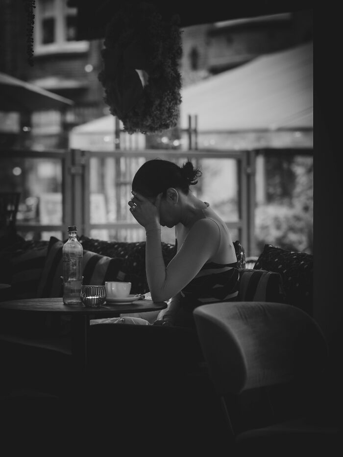 Bartenders overhear: a woman at a cafe table, head in hands, looking upset, possibly from a overheard conversation.
