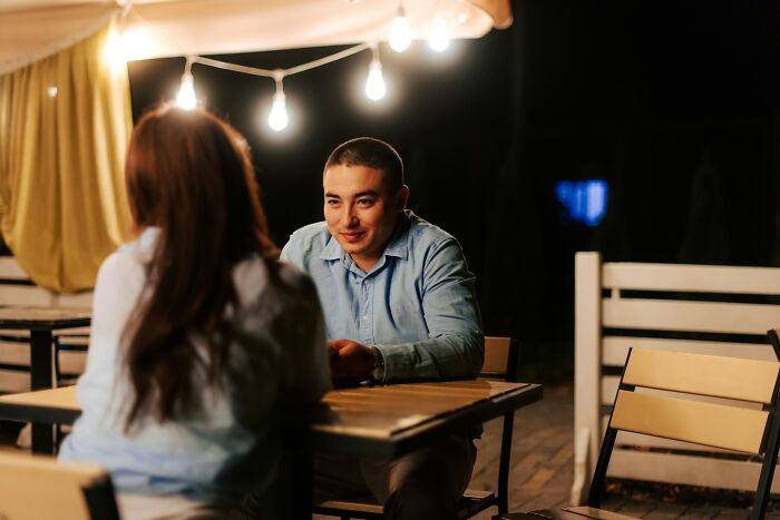 A man smiling at a woman across a table outdoors at night, with conversations overheard by bartenders.