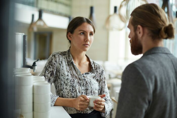 Bartenders overhear: two people engaged in a serious conversation at a cafe counter, one holding a cup.