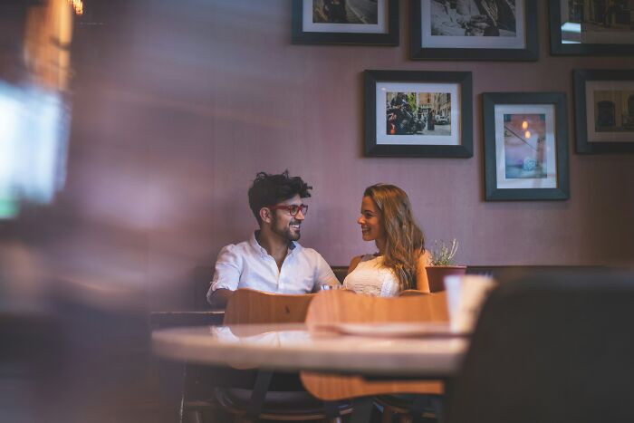 A couple smiling at each other in a dimly lit restaurant, enjoying their conversation overheard by bartenders.