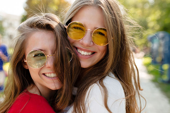 Two young women with long hair and round sunglasses smiling at the camera, conveying friendship and a potential bridesmaid role. Two young women with long hair and round sunglasses smiling at the camera, conveying friendship and a potential bridesmaid role.