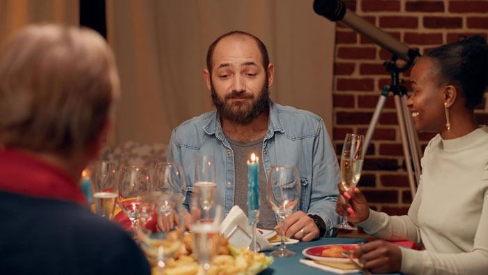 A man with a beard sits at a dinner table looking uncomfortable, a GF claims she hasn't spoken to him in years.