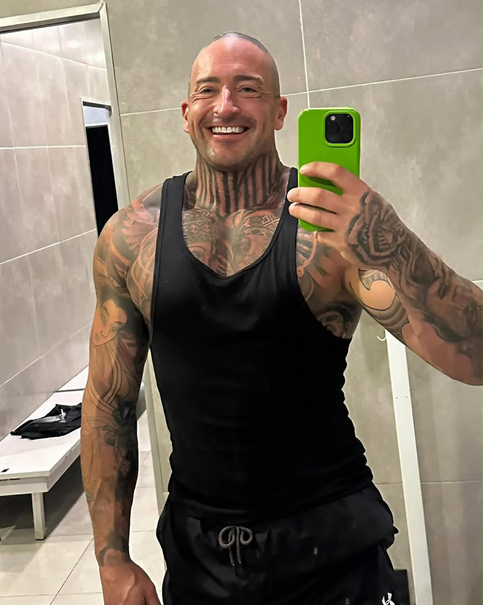 A heavily tattooed man in a gym, smiling while taking a selfie, relevant to Bonnie Blue's coworker spilling tea.
