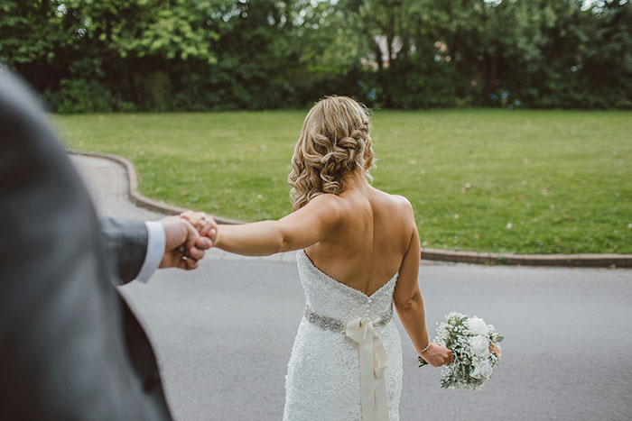Back of a bride in a white wedding gown, holding a bouquet and reaching for the groom's hand, representing twisted values.