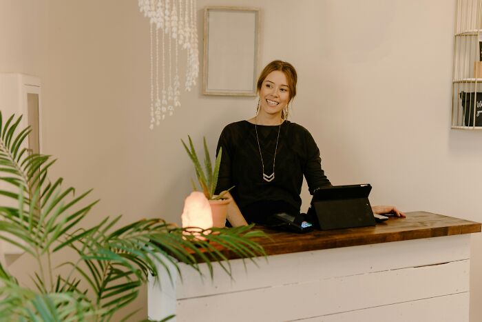 A smiling woman behind a wooden counter with a tablet, plants, and a salt lamp, depicting a welcoming office environment.