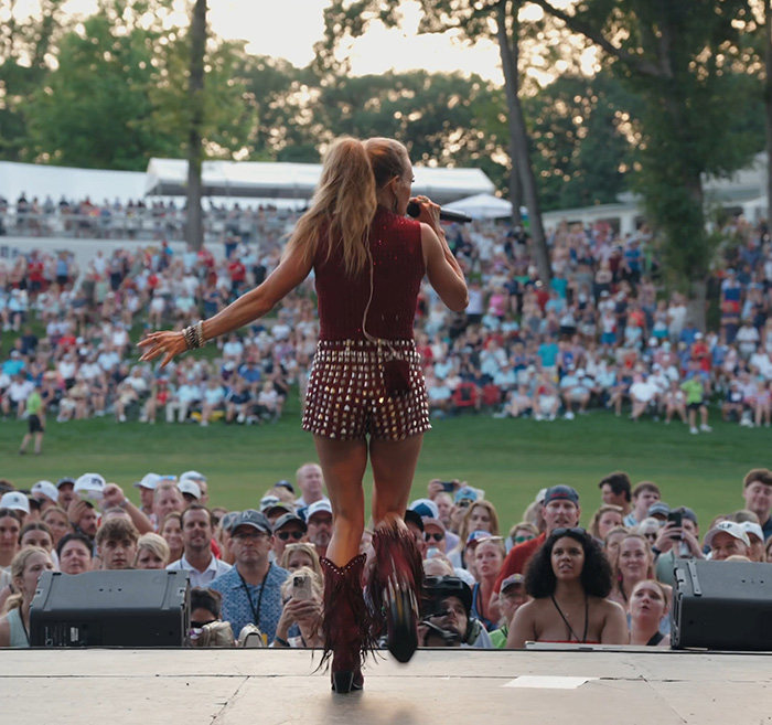 Carrie Underwood performing on stage at a golf course, surrounded by a large crowd.