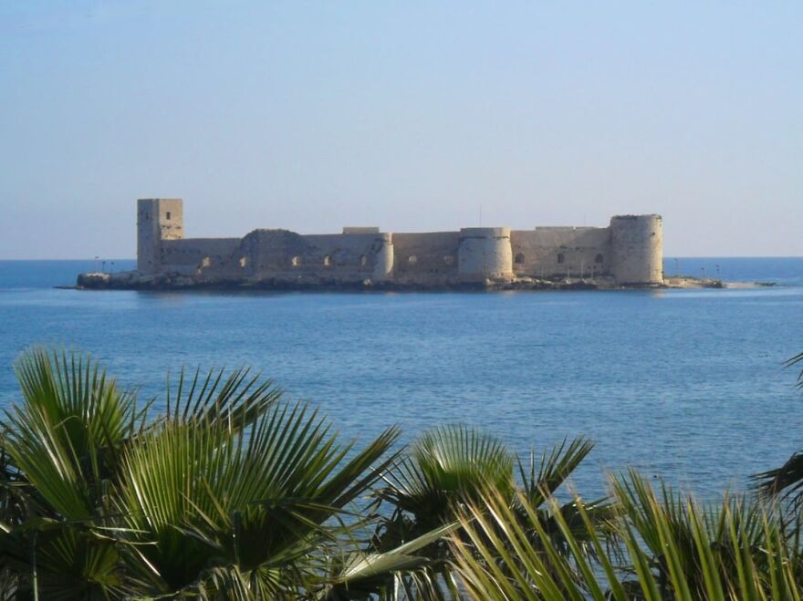 Beautiful and interesting island castle in the middle of a calm blue sea, framed by palm fronds.