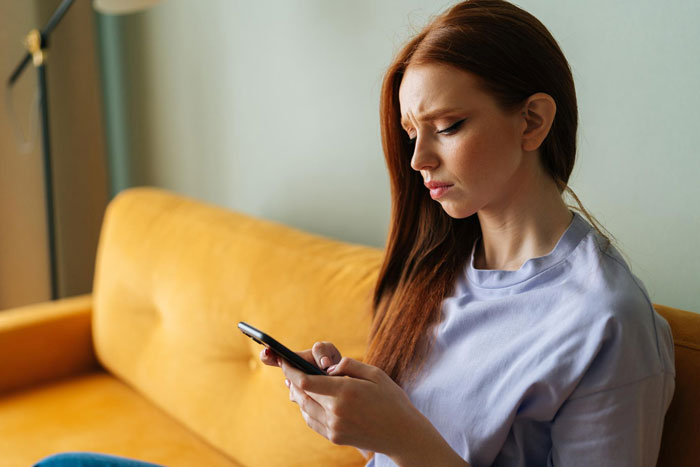 A red-haired woman on a yellow couch looking confused at her phone, depicting a bride whose bestie ghosts her on wedding day. A red-haired woman on a yellow couch looking confused at her phone, depicting a bride whose bestie ghosts her on wedding day.
