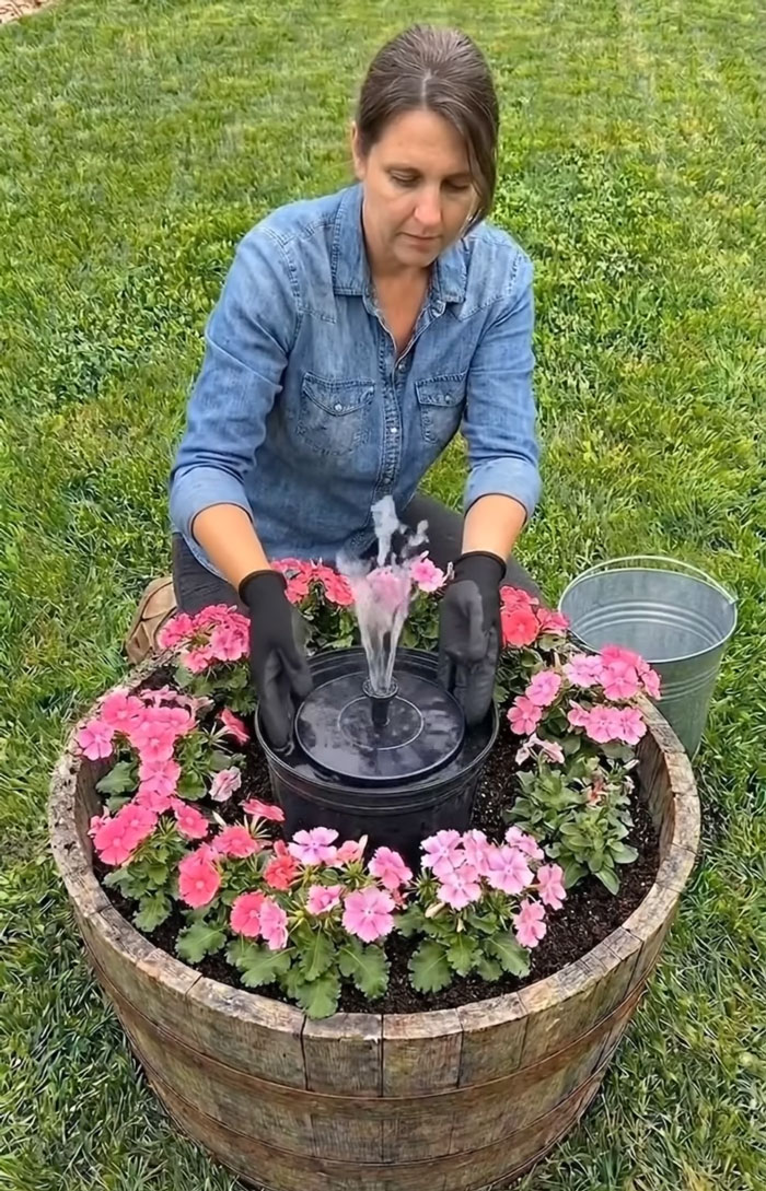 Woman installing a small water fountain surrounded by pink flowers in a wooden barrel, showcasing clever craft projects.