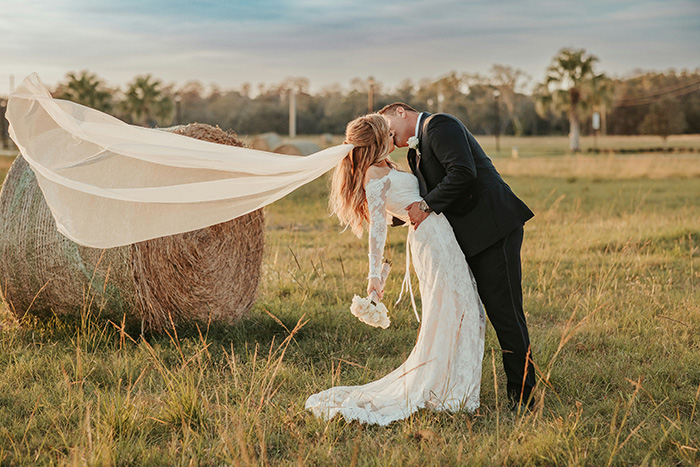 A newlywed couple shares a kiss in a field with a hay bale, veil blowing in the wind during a wedding. A newlywed couple shares a kiss in a field with a hay bale, veil blowing in the wind during a wedding.