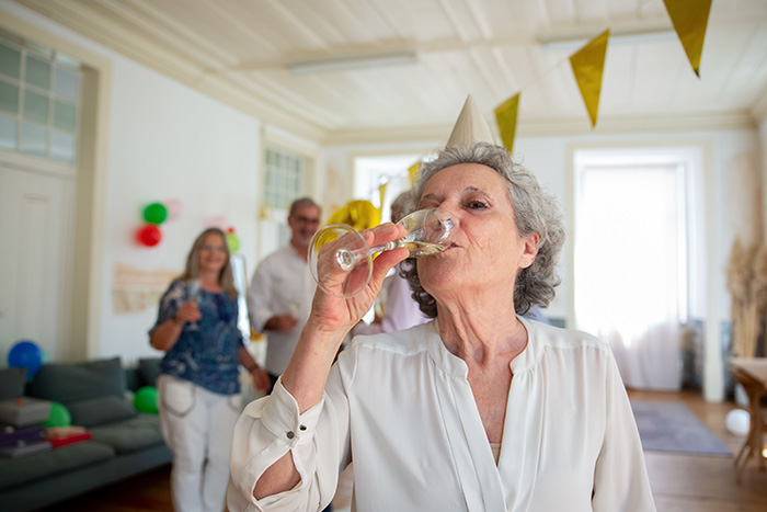 A MIL at a wedding in a party hat drinks from a champagne flute, guests in the background at a decorated venue. A MIL at a wedding in a party hat drinks from a champagne flute, guests in the background at a decorated venue.