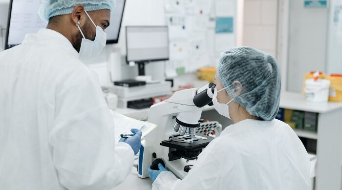 Two scientists in a lab, one looking into a microscope and the other taking notes, studying human body biology.