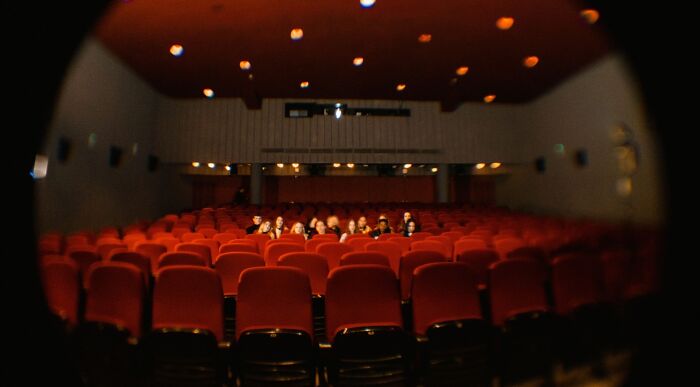 A wide shot of a classic movie theater with red seats, a few people are scattered throughout the rows. A movie expert can identify the actors. A wide shot of a classic movie theater with red seats, a few people are scattered throughout the rows. A movie expert can identify the actors.