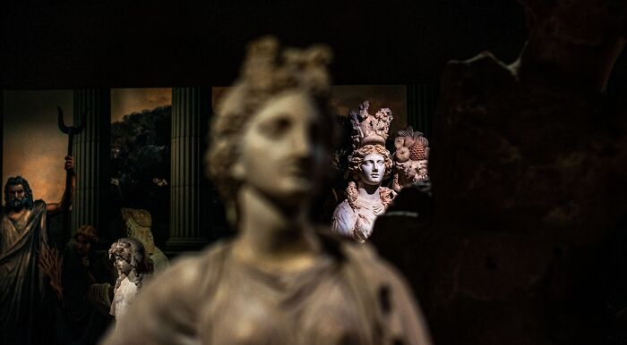 A close-up shot of Greek mythology statues, with one in the foreground blurred and others in the background in focus. The statues are made of white marble and depict human-like figures with intricate details, including flowing hair and robes. In the far background, a painting of ancient Greek figures is visible between two dark columns. The overall lighting is dim, highlighting the statues. This image represents the Greek mythology quiz. A close-up shot of Greek mythology statues, with one in the foreground blurred and others in the background in focus. The statues are made of white marble and depict human-like figures with intricate details, including flowing hair and robes. In the far background, a painting of ancient Greek figures is visible between two dark columns. The overall lighting is dim, highlighting the statues. This image represents the Greek mythology quiz.