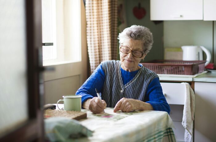 An elderly woman with glasses writing at a table, representing the act of creating a Last Will.
