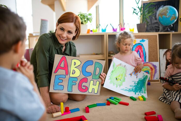 A happy teacher with red hair shows a sign with ABCDEFGH to two children in a colorful classroom, with funny last will stories.
