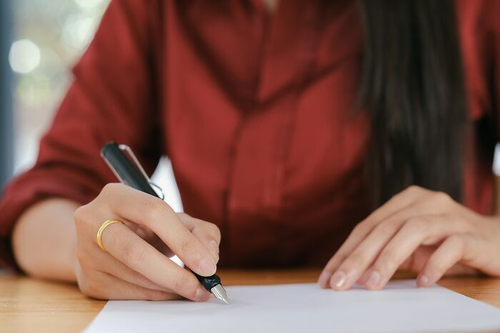 A close-up of a person's hand with a gold ring writing on a blank paper, possibly for a serious last will story.