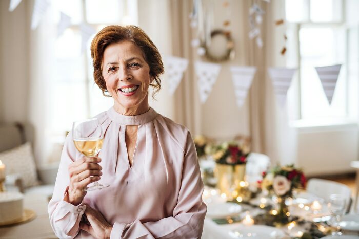 A smiling woman holding a glass of white wine in a decorated room with a festive table in the background. This is not about last will stories.