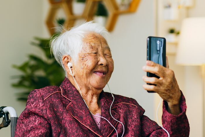 An elderly woman smiling while on a video call with earbuds, showing connection and memories for Last Will Stories.