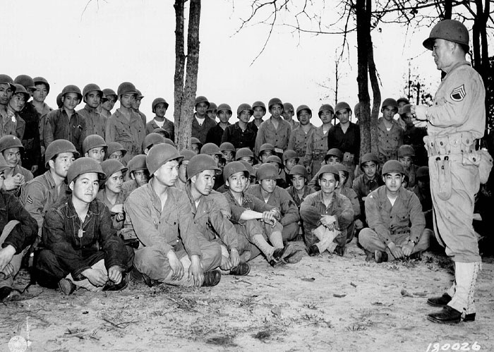 America: Soldiers in uniform and helmets, seated on the ground, listening to an instructor.